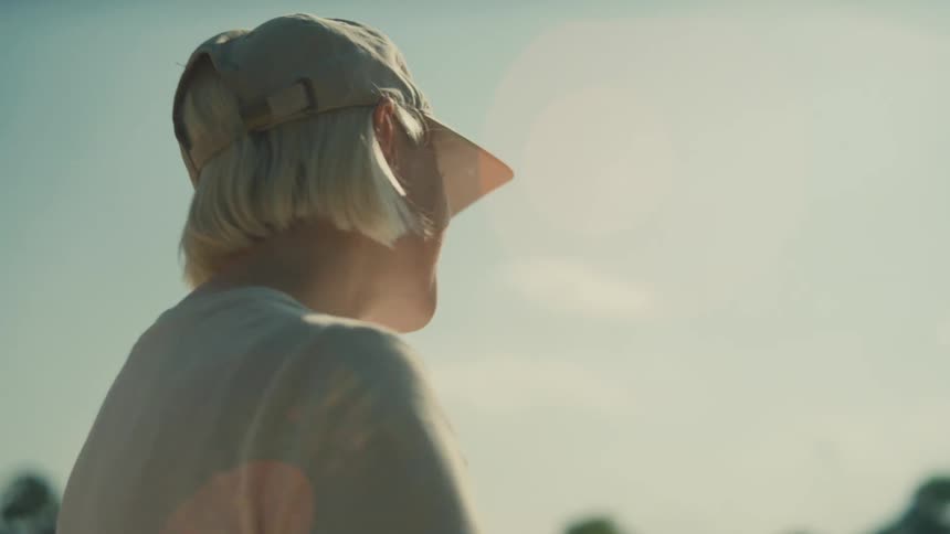 A person with short blonde hair and a baseball cap waves to someone off screen under a bright sunny sky with lens flare