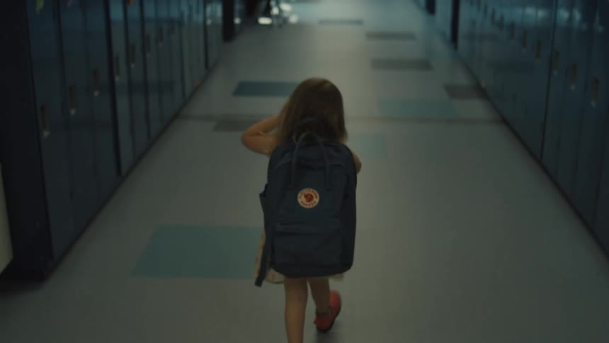 A little girl with light brown hair and a dark blue backpack is walking down an empty school hallway lined with blue lockers