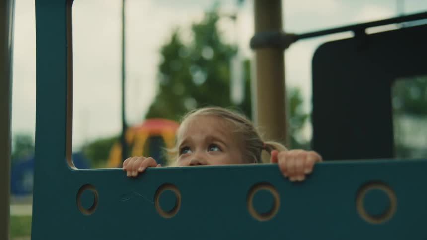 A small girl with blonde pigtails is playing on a playground she is wearing a blue dress and a small blue backpack with a red fox emblem on it she happily talks to an adult offcamera