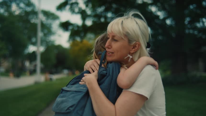 A mother and daughter are walking together on a sidewalk the mother kneels down to hug the little girl who is wearing a backpack and a dress