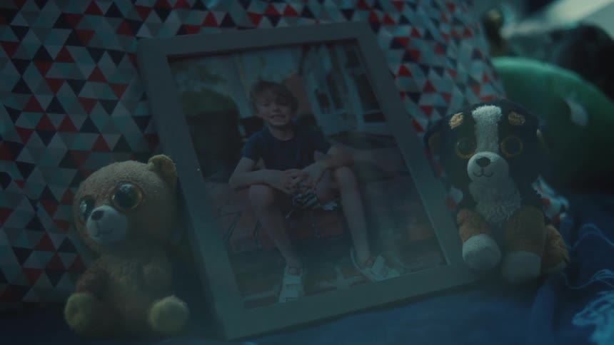 Framed picture showing a smiling boy sits on a bed between stuffed toys