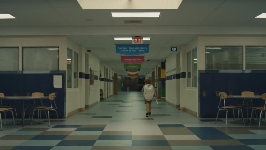 A young boy wearing a backpack walks down an empty school hallway the hallway has rows of lockers tables and chairs and a checkered tile floor he walks away from the camera and down the long corridor