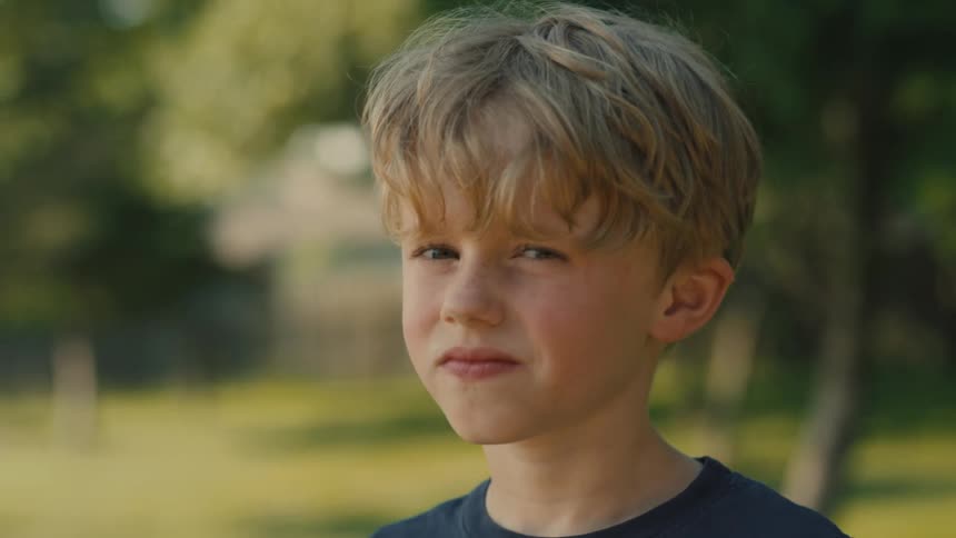 A young boy with blonde hair and a blue shirt looks directly at the camera with a neutral expression blinking slowly with a blurred outdoor background