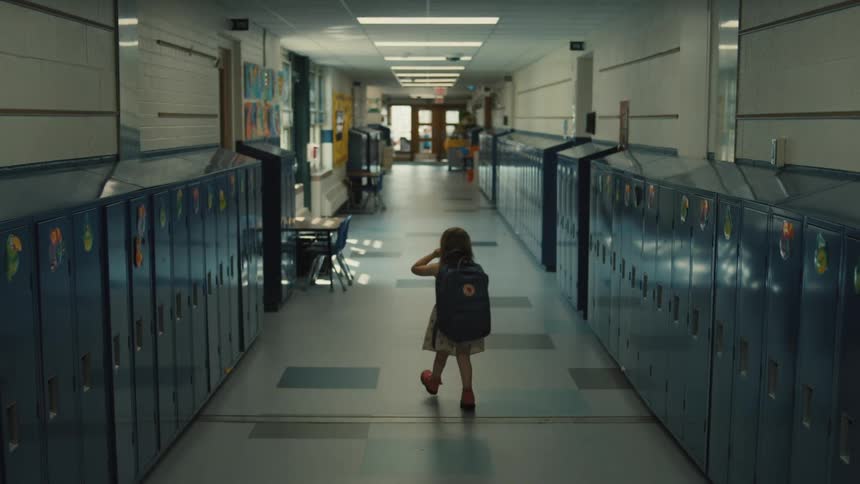 A child with a backpack walking alone down a long empty school hallway lined with lockers