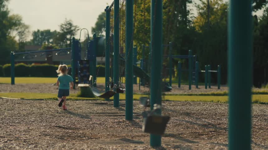 The video shows a playground on a sunny day a young boy and a young girl are swinging on swings while another child runs past them in the background