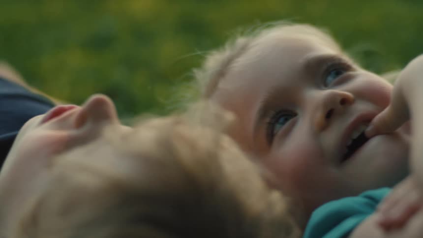 The video shows two young children lying on the grass together looking up one child is smiling and has their finger in their mouth