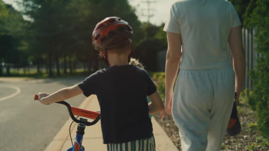 A child wearing a helmet pushes a bicycle while walking alongside another child and an adult on a sunny sidewalk