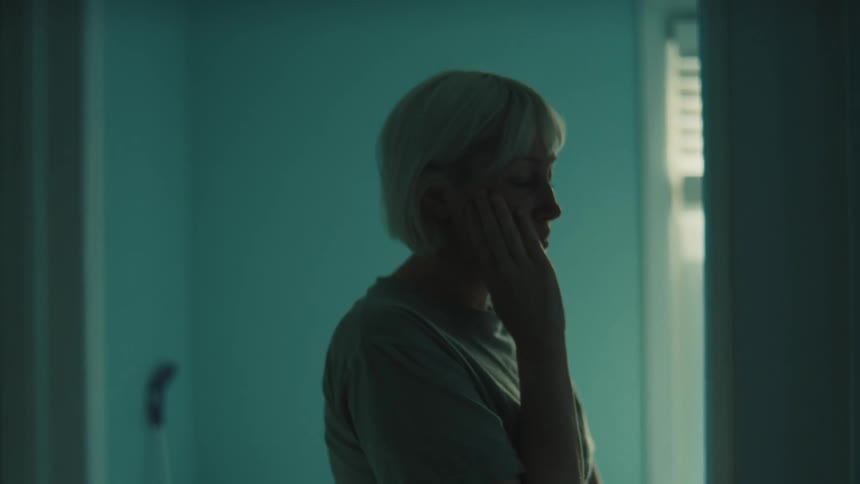 A young woman stands in a doorway indoors facing the wall with her hands on her cheeks looking sad