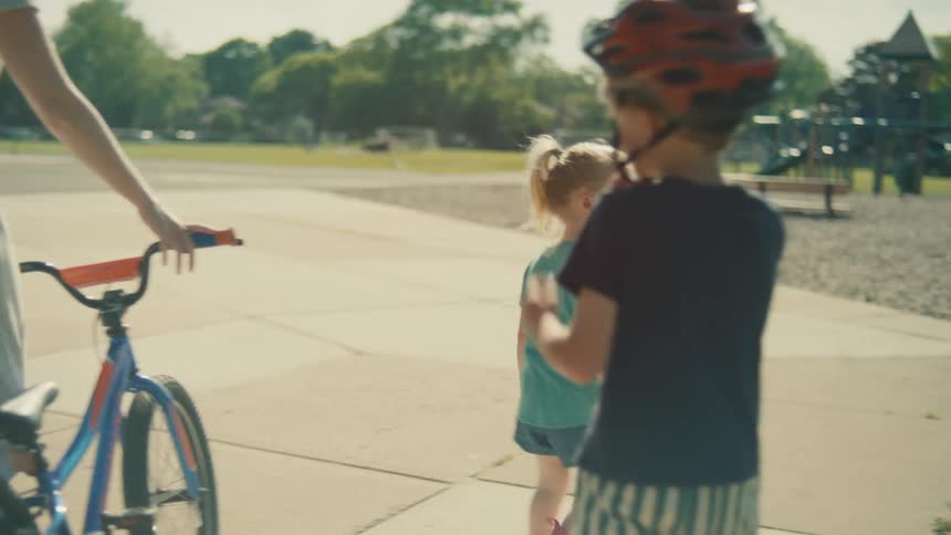 Mother accompanies two children as one adjusts the helmet next to the bicycle