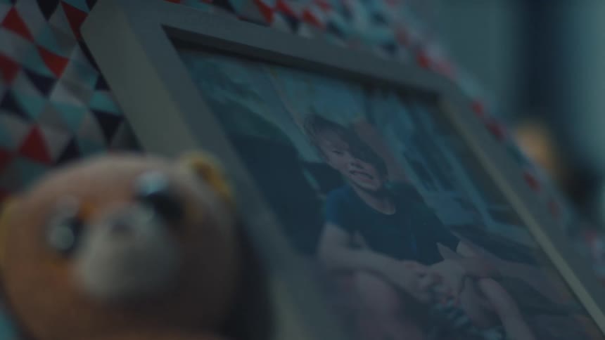 A close shallow focus view shows a framed picture of a young boy with light colored hair and a blue shirt sitting with his arms resting on his knees while a blurry brown teddy bear is visible in the foreground