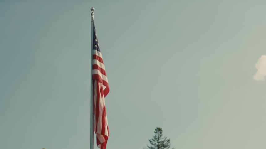 The video shows a lowangle shot of an american flag on a flagpole against a clear sky the flag is hanging still with no wind blowing