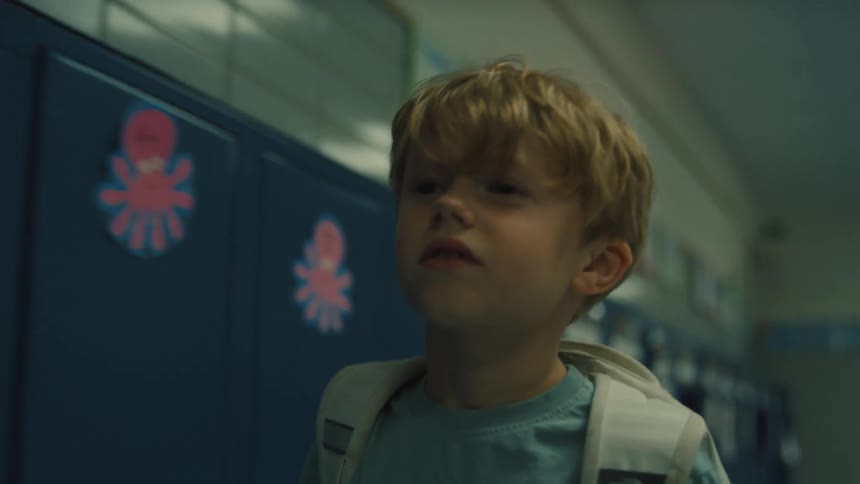 A young boy walking with his chin up on a school hallway his hand running through a line of blue lockers all with individual childrens art on them