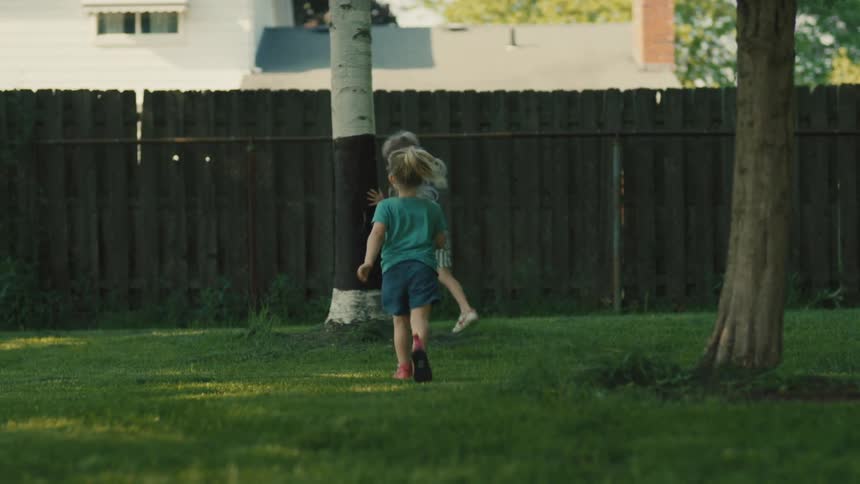 Two kids play in the backyard lawn at midday chasing each other around a tree