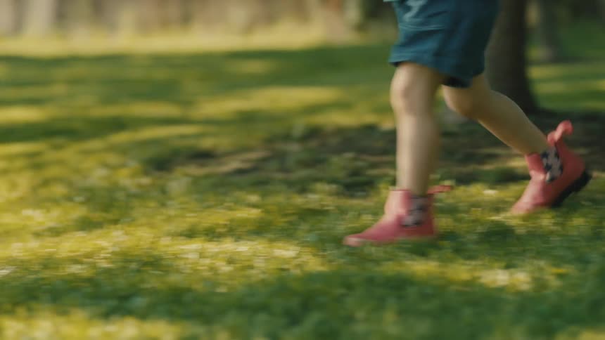 A closeup of a young childs legs and red shoes as they walk and then run playfully across a green sunlit lawn