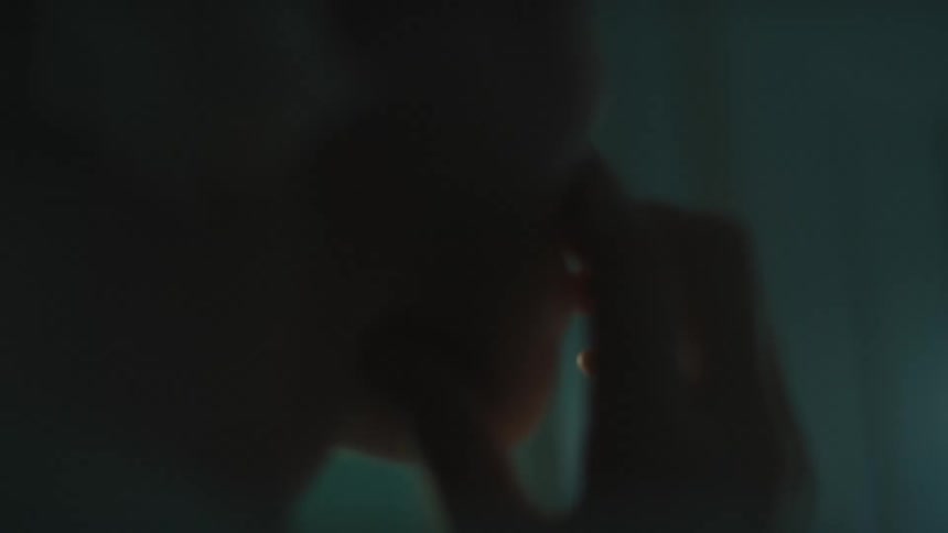 A blonde haired individual touches their face with a melancholic expression looking out of frame in a dimly lit setting