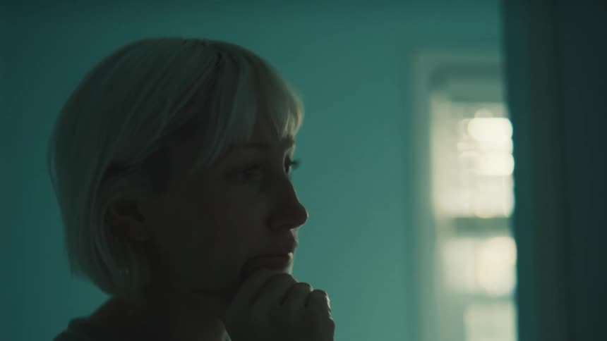 A side profile closeup of a woman with short blonde hair standing in a doorway with her hand on her chin