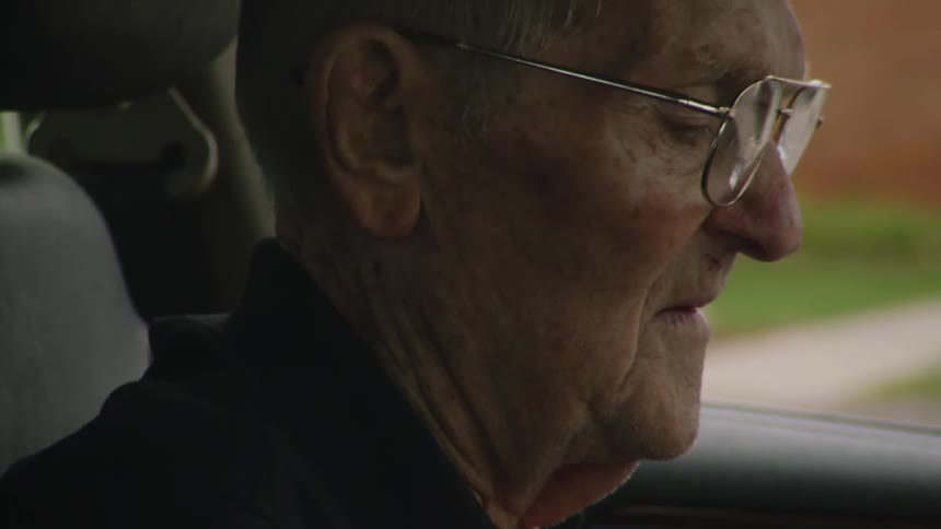 The video shows an elderly man with glasses in the passenger seat of a car he is holding a flip phone and appears to be smoking a cigarette