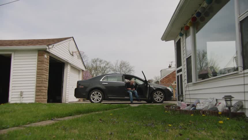 A person sits in the drivers seat of a dark sedan with the door open parked in a suburban driveway next to a garage and a house