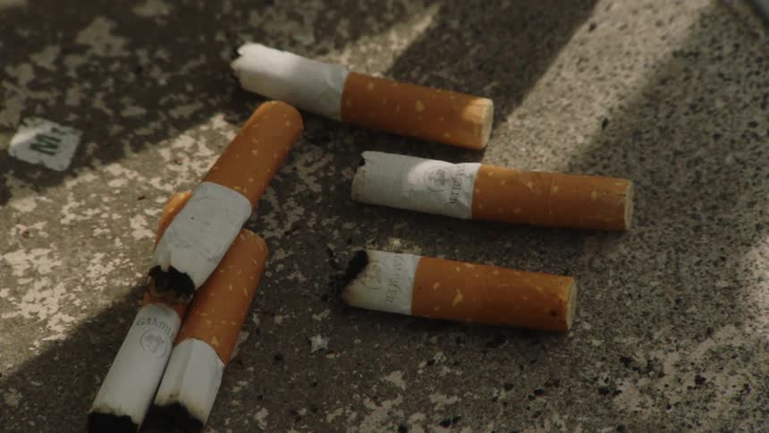 A close view of a dirty gray tabletop shows several cigarette butts with brown filters and white paper scattered across the surface