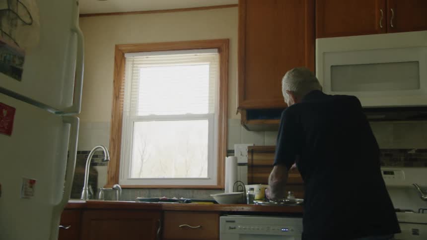 An older man seen from behind stands in a brightly lit home kitchen and uses a spatula to serve freshly cooked food from a pan onto a plate