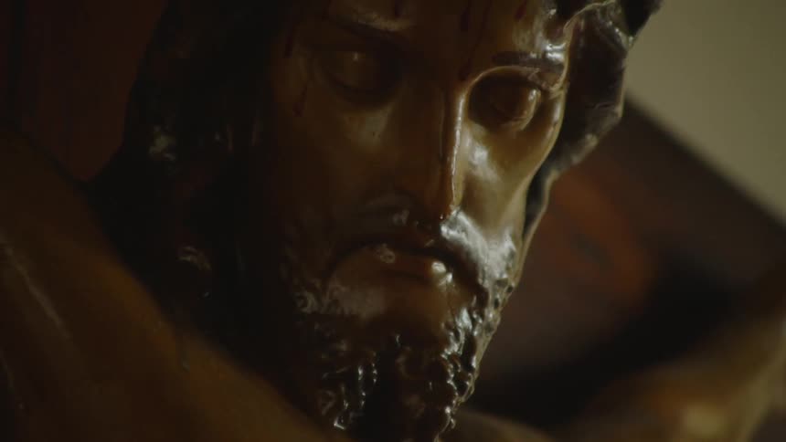 A close low angle view shows the face and right arm of a wooden statue of jesus on a cross with a reflective glossy finish that catches the light