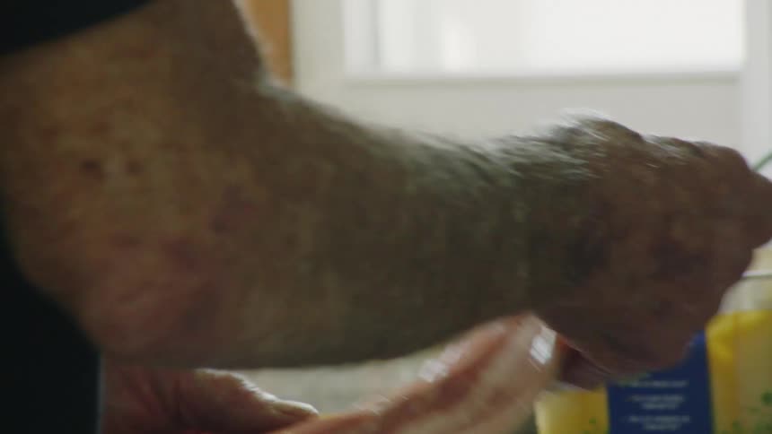 An elderly persons hands are shown preparing bacon in a kitchen with a container of butter and other kitchen items in the background