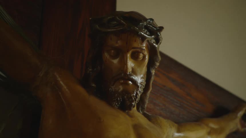 A closeup shot of a wooden crucifix focusing on the carved figure of jesus christ before slowly zooming out to reveal more of the religious sculpture