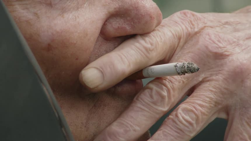 An elderly man sits in the passenger seat of a car at midday facing outside while smoking a cigarette
