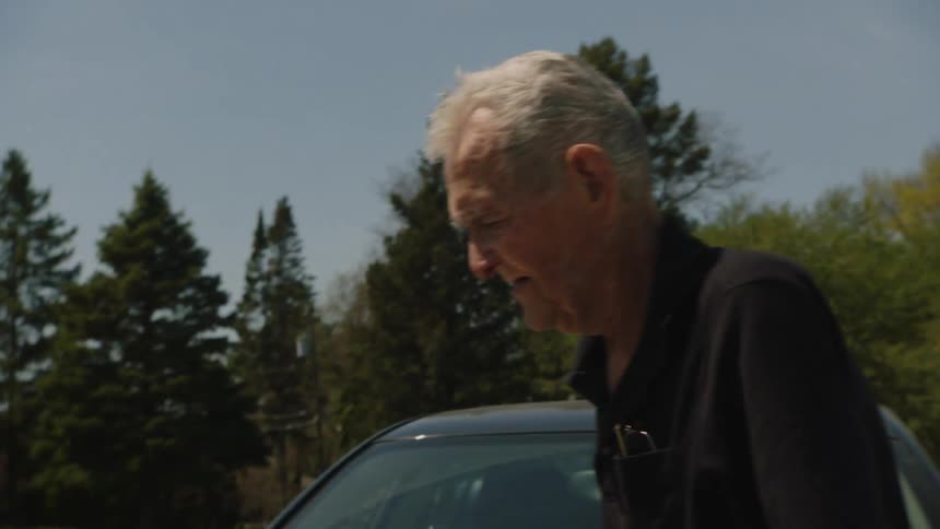 The video shows a man with white hair standing next to a car with an open door he is holding a cigarette and looking at the camera he then begins to walk away from the camera using a cane