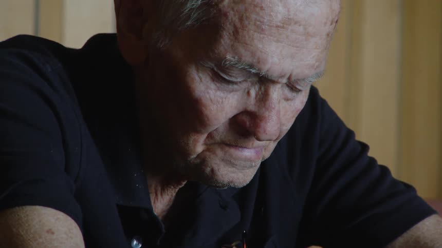 The video shows an elderly man sitting at a table with his hands clasped together looking down he has short white hair and is wearing a dark polo shirt