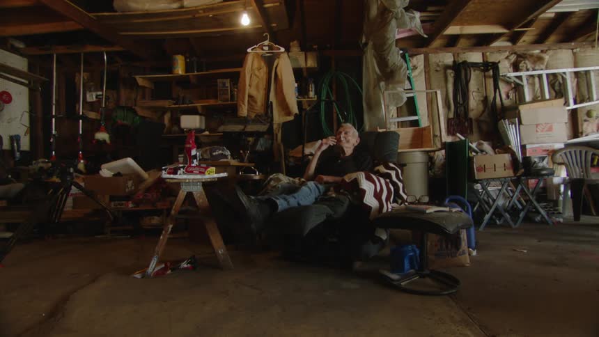 An elderly man sits in a recliner in a cluttered garage with various items like tools shelves and a ladder visible in the background