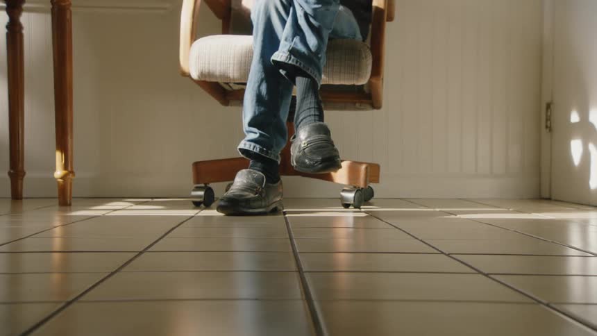 A man wearing blue jeans and black shoes is sitting with his legs crossed on a rolling office chair slowly moving his feet as sunlight streams in from the right