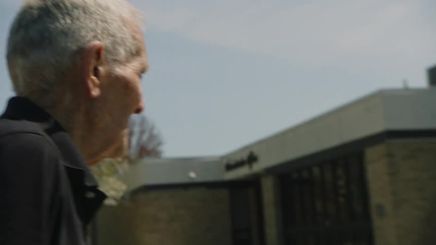 A man with white hair and a black collared shirt is walking on a sidewalk smoking a cigarette and holding it in his right hand