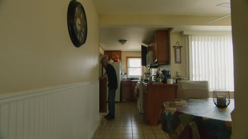 An elderly man walks around the kitchen in a quiet home setting
