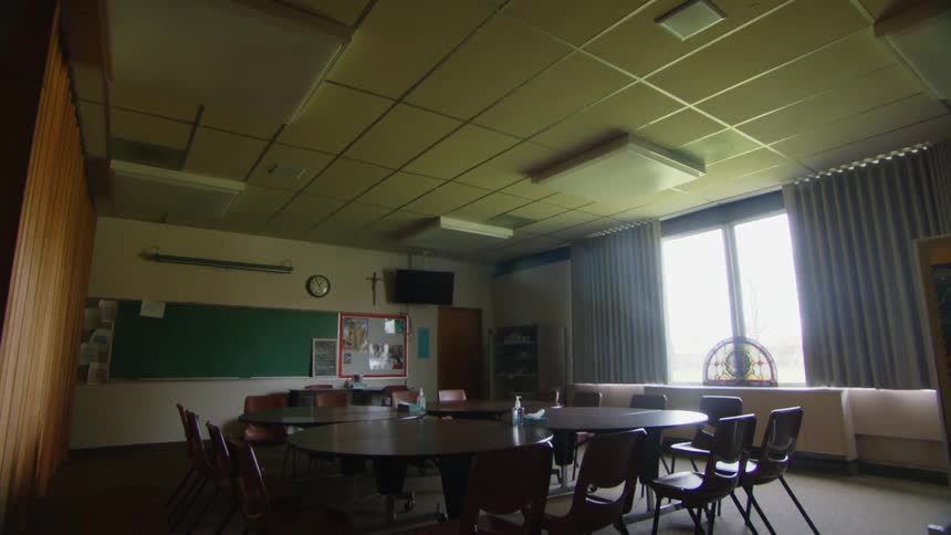 A camera begins by looking at a ceiling with fluorescent lights then pans down to show a classroom with round wooden tables surrounded by brown plastic chairs and a chalkboard on the left wall with a television hanging above it