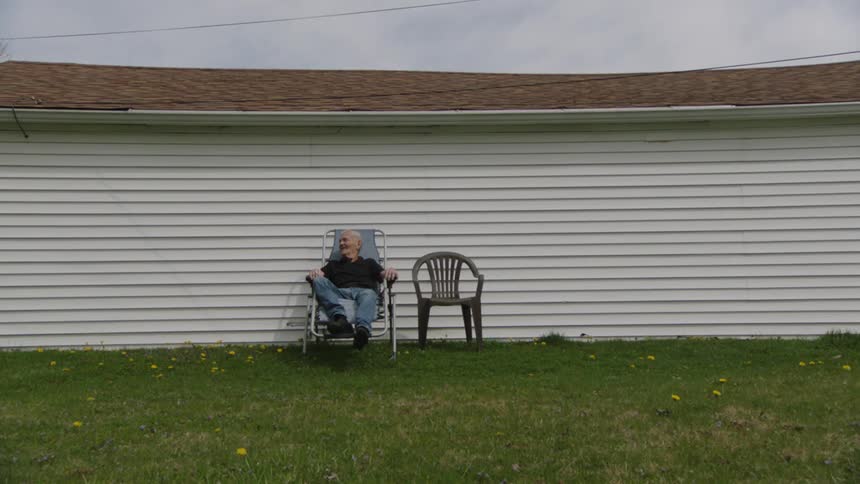 The video shows a man with white hair wearing a dark polo shirt and blue jeans sitting in a folding chair on a grassy lawn he is relaxing and smoking a cigarette a white building with horizontal siding is in the background