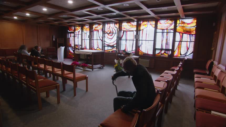 A person with white hair and glasses sits on a chair in a room with stained glass windows and rows of empty seats holding a cane