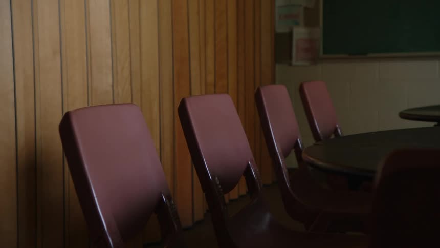 A long view shows a dark room with a green blackboard and a long table with four chairs lined up on its left side against a wood paneled wall