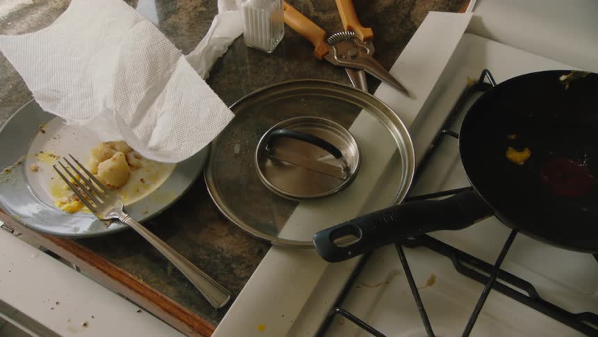 A dirty and messy kitchen counter with a dirty pan on the stove and a dirty dish with leftover potatoes on the counter