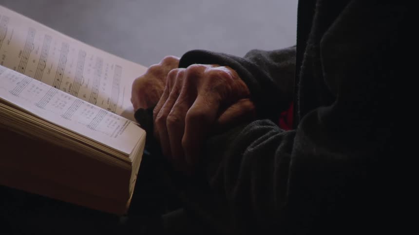 An elderly person clasps their hands over an open hymn book