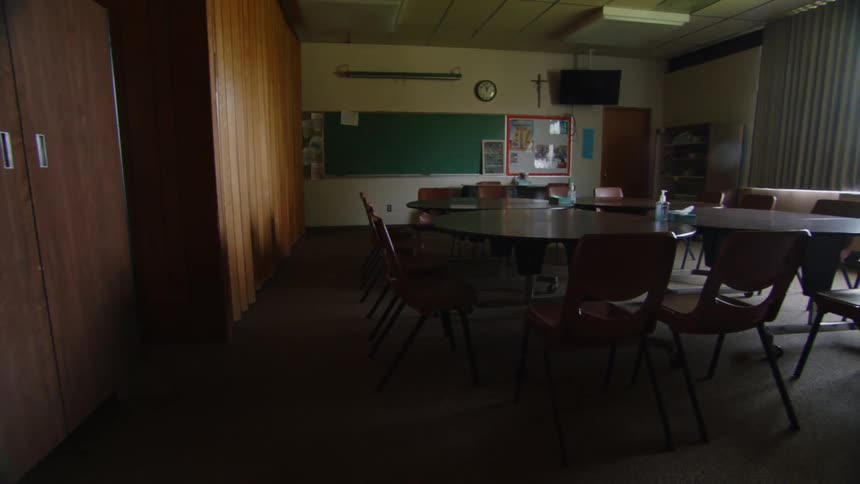 The video shows an empty dimly lit classroom there are several large round tables with chairs and a green chalkboard at the front the room has a retro feel with dark wood cabinets and vertical blinds on a large window