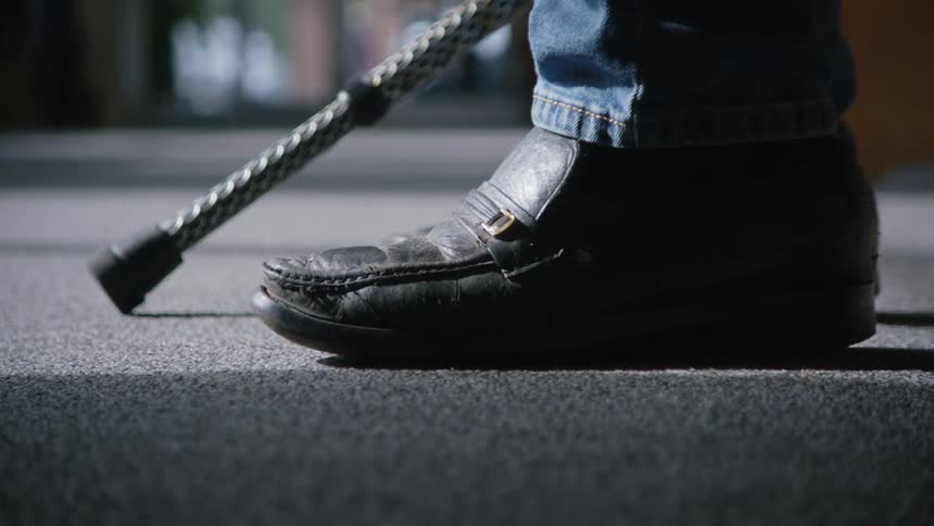 A closeup shot of an elderly mans black shoes and the base of his walking cane