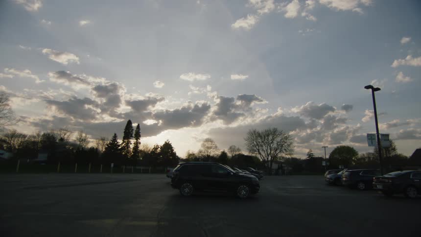 The video shows a wide shot of an empty parking lot on a cloudy day with a few cars parked a black suv drives slowly across the lot and exits the frame