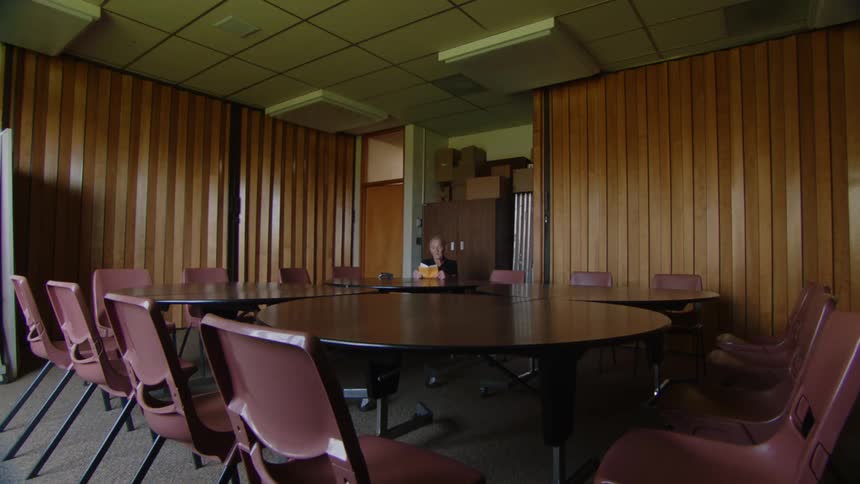 An elderly man sits alone reading a book at the far table of a large dimly lit and dated conference room with woodpaneled walls and empty chairs