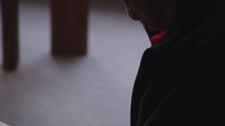 A closeup shot shows a persons hands holding an open book with sheet music the person is wearing a dark hoodie and glasses and is looking down at the book
