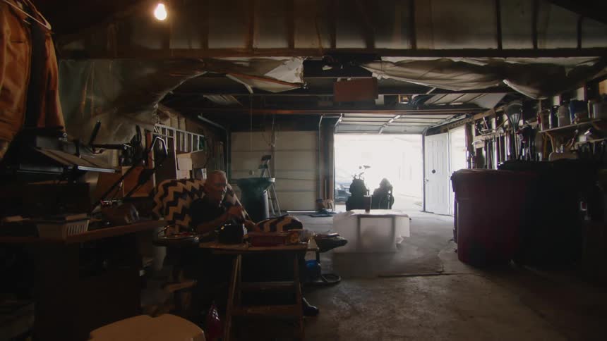 An elderly man sits in a dimly lit garage smoking a cigarette