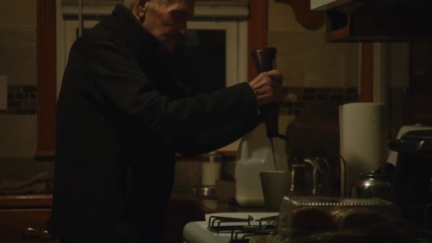 As the camera slowly pushes in an elderly man in a dark jacket stands in a dimly lit kitchen and carefully squeezes chocolate syrup from a bottle of a bottle into a mug