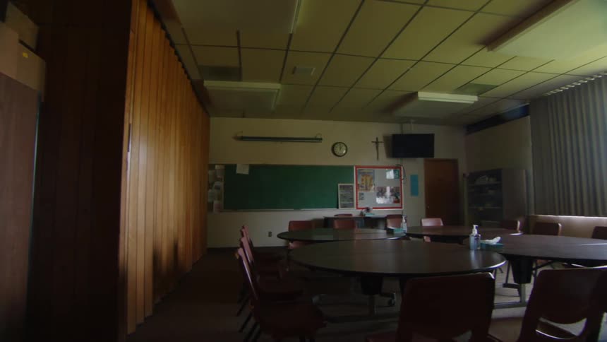 A slow panning shot reveals an old dated and empty meeting room in a church with chairs stacked tables in disarray and a cross still hanging on the wall