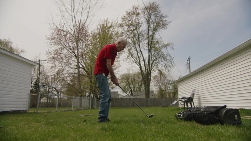 An elderly man practices golf swings in a suburban backyard