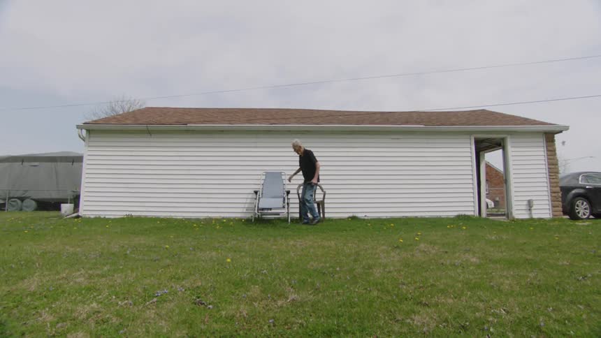 An elderly man walks behind his house and sits in a chair